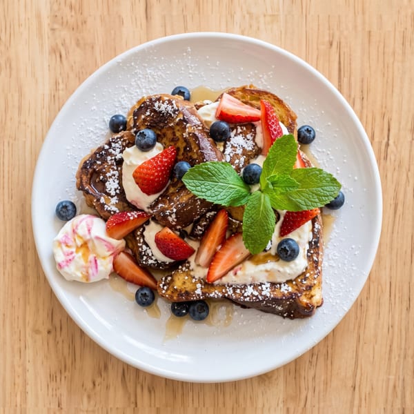 French toast with strawberries, blueberries, whipped cream, powdered sugar, and mint on a white plate.