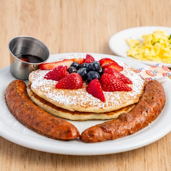 Pancakes with strawberries, blueberries, powdered sugar, sausage links, syrup, and scrambled eggs.