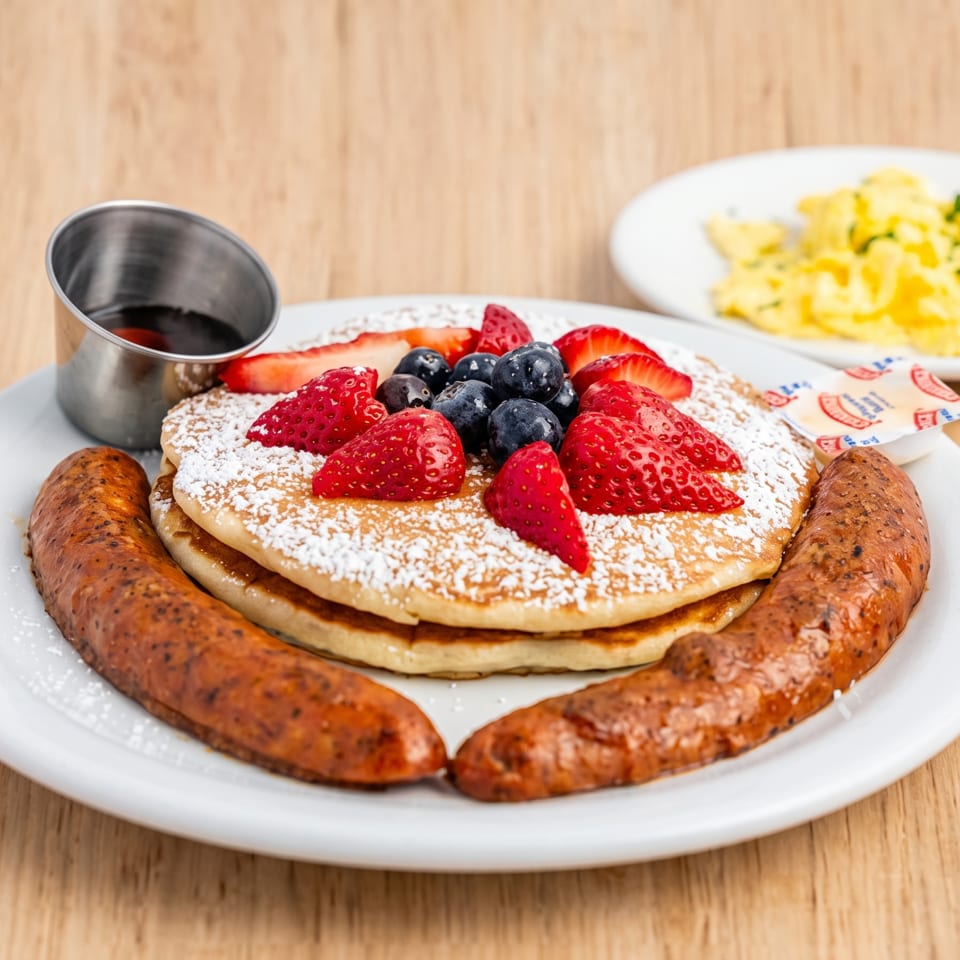 Pancakes with strawberries, blueberries, powdered sugar, sausage links, syrup, and scrambled eggs.